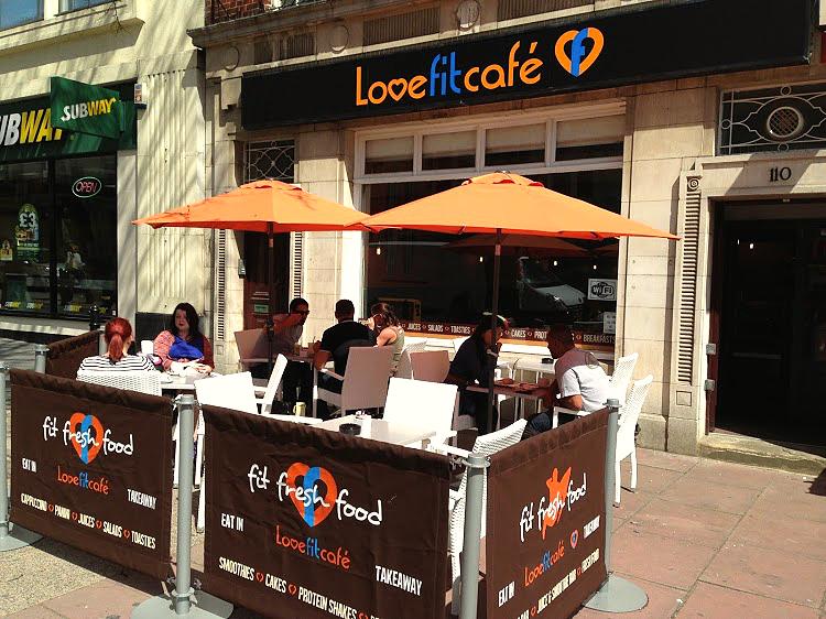 Exterior of Lovefit Café with black and orange signage. Customers sit outside under bright orange parasols, enjoying food and drinks. The café promotes fit and fresh food, located next to a Subway branch on a busy street.