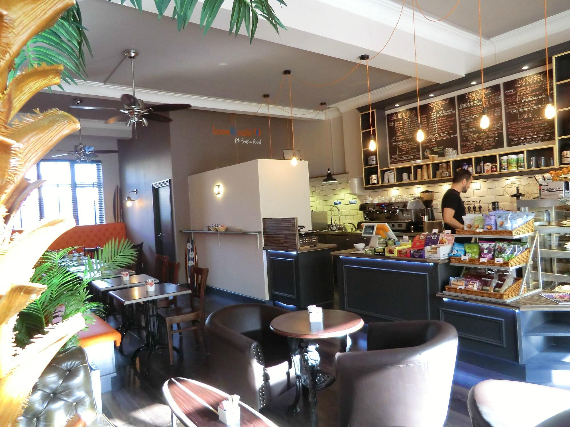 Modern café interior with a view of the counter, where a barista prepares drinks. Chalkboard menus hang above the coffee machines, and the space features hanging filament lights, dark wood floors, and relaxed seating with leather armchairs and small tables.